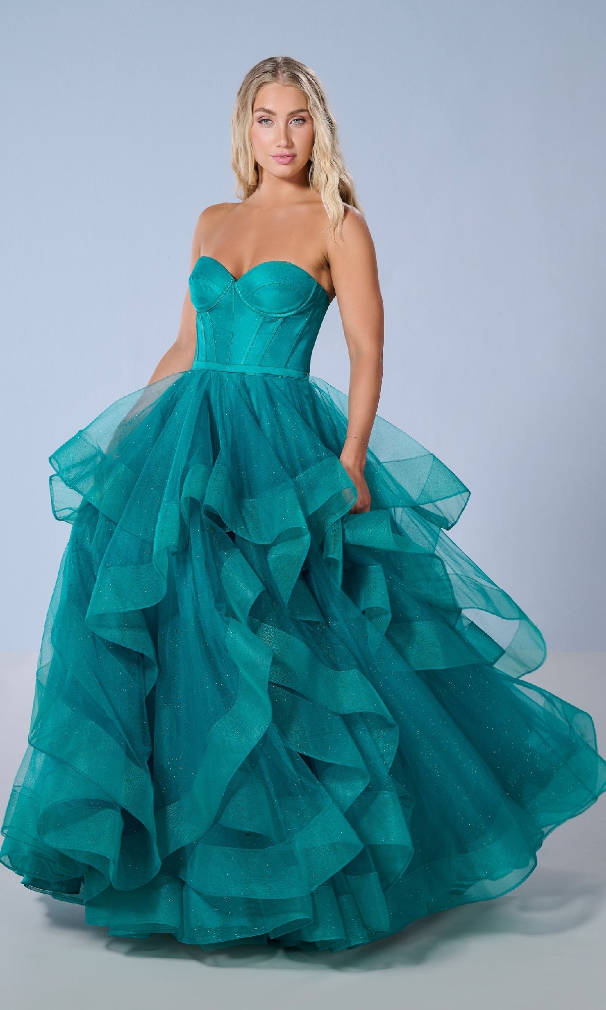 A woman wearing the Tiffany 16245 long prom dress, featuring a strapless corset bodice and layered ruffled glitter tulle skirt, stands against a plain light blue background.